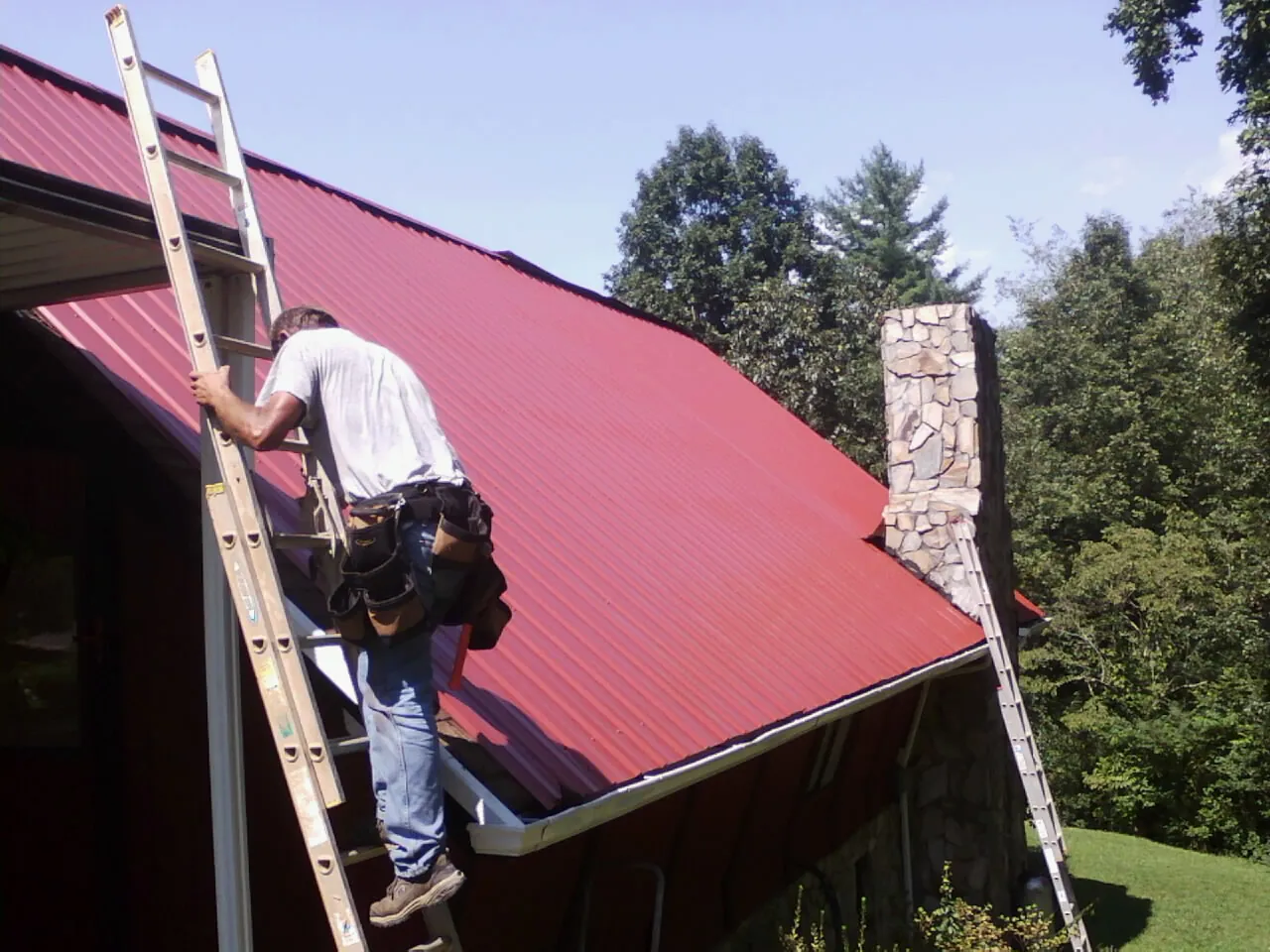 Roofer on a ladder beside a red metal roof and stone chimney — Soffit & Fascia Repair by Roofing Brothers in Hoffman Estates